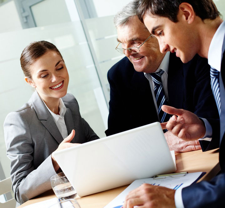 Three business professionals dressed in suits gathered around a desk, smiling and engaging in discussion while looking at a computer screen.