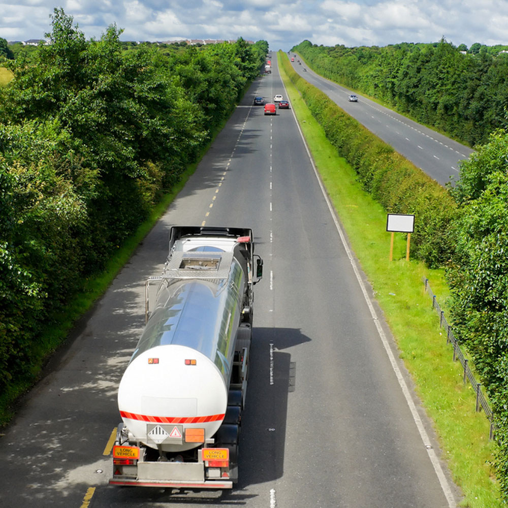 Aerial view of a tank truck driving on a straight road through the countryside, with other cars visible in the distance.