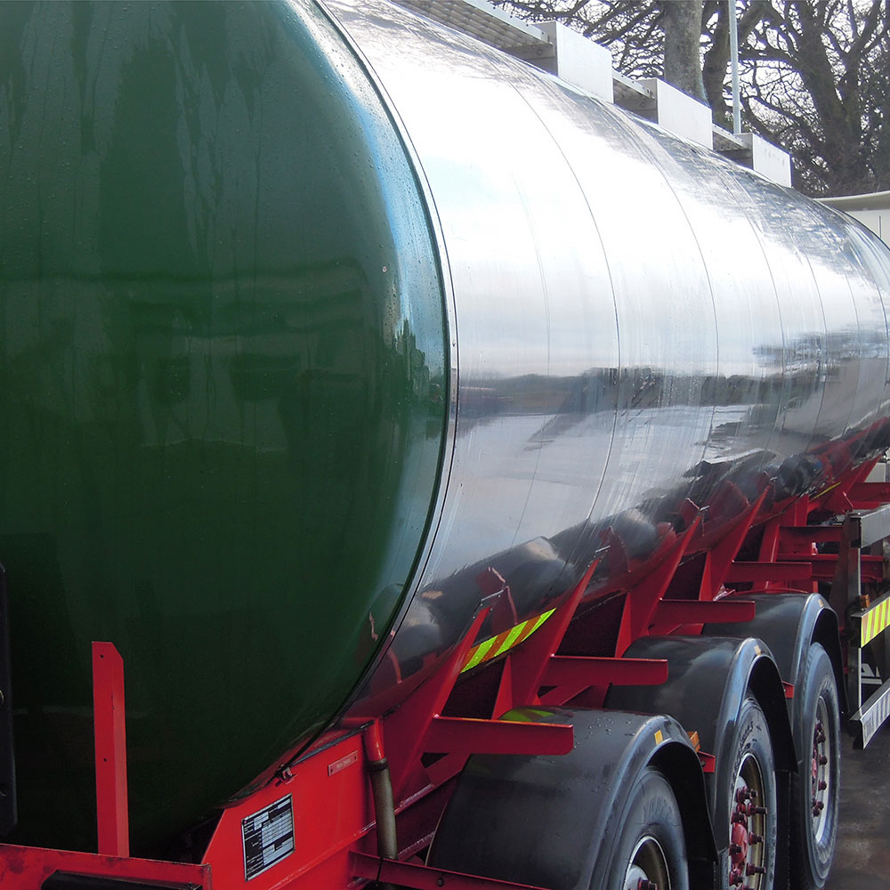 A shiny tank truck parked outdoors, reflecting the road on its polished surface. Three visible wheels are secured with red metal braces.